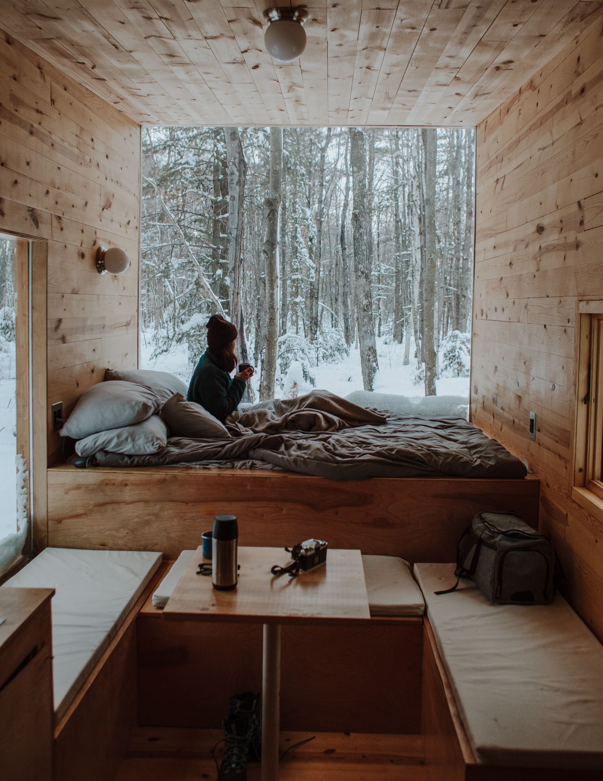 a woman huddled in bed with blankets and a jacket looking out a window to a wintery view